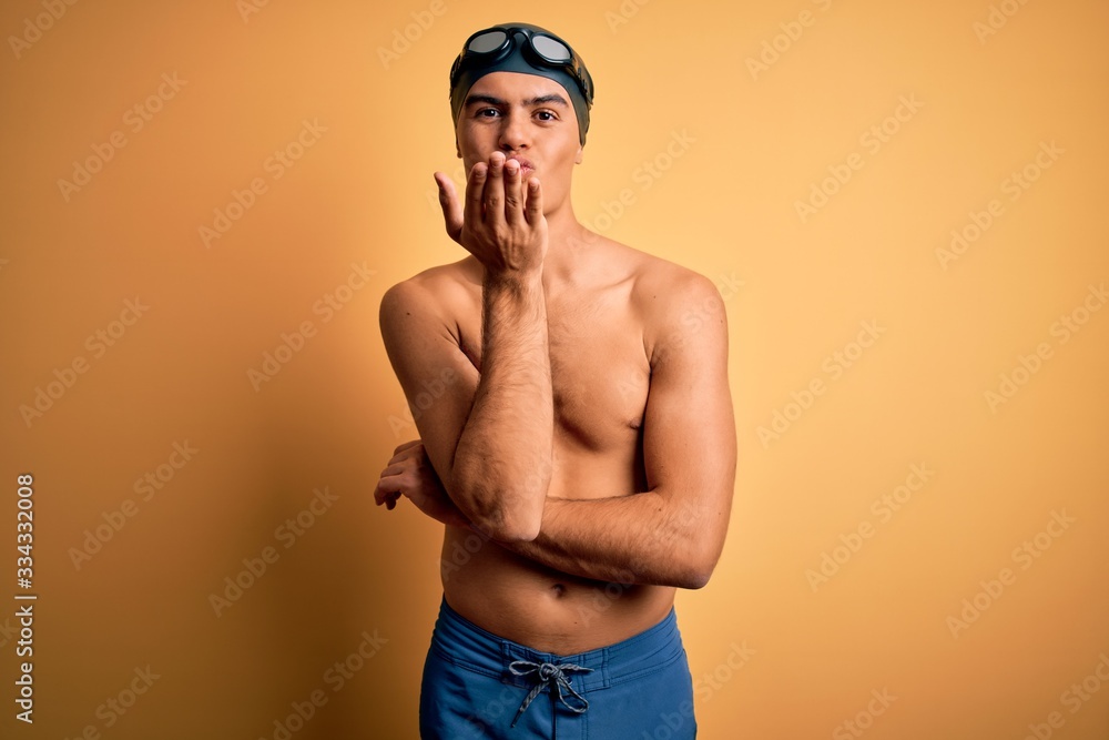 Young handsome man shirtless wearing swimsuit and swim cap over isolated yellow background looking at the camera blowing a kiss with hand on air being lovely and sexy. Love expression.