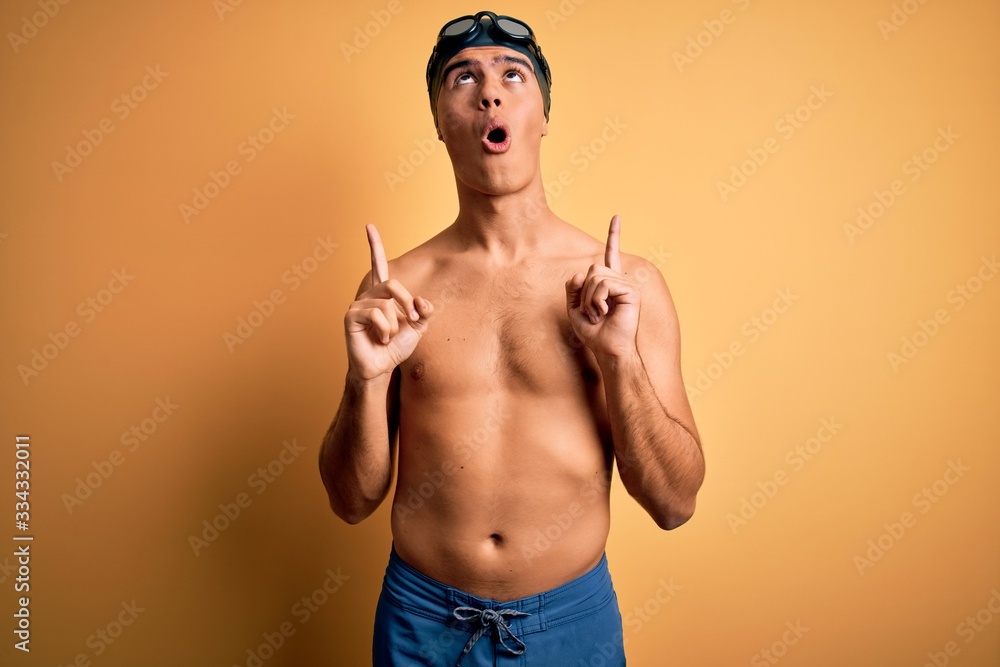 Young handsome man shirtless wearing swimsuit and swim cap over isolated yellow background amazed and surprised looking up and pointing with fingers and raised arms.