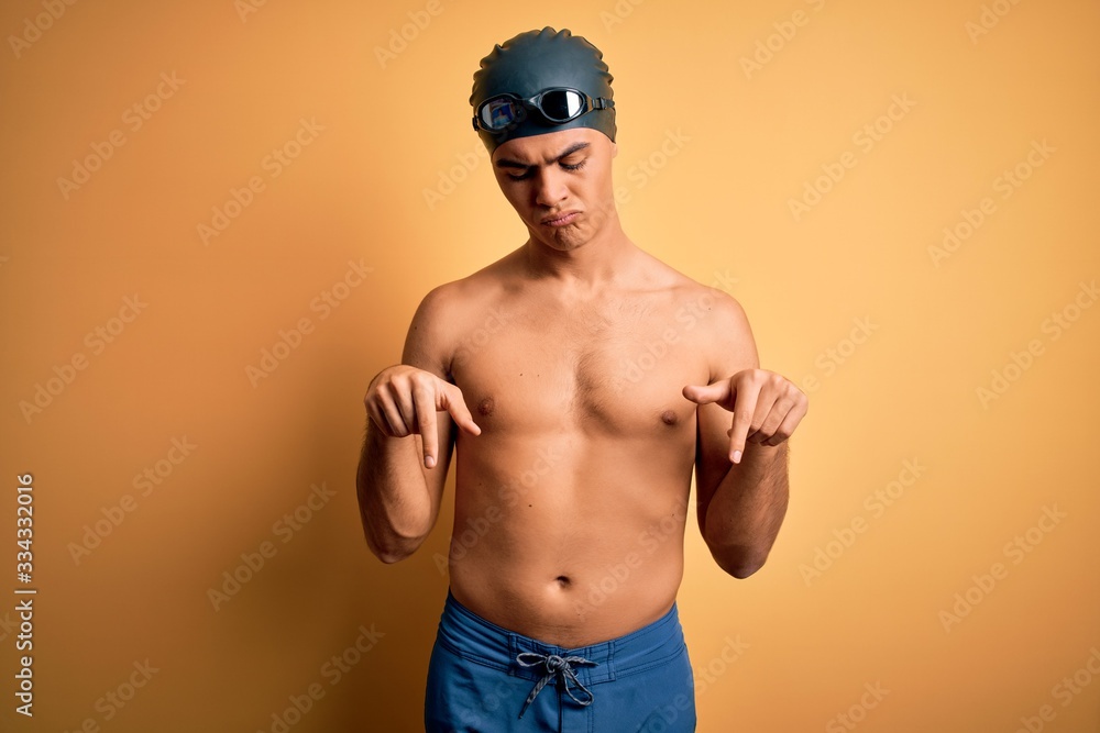 Young handsome man shirtless wearing swimsuit and swim cap over isolated yellow background Pointing down looking sad and upset, indicating direction with fingers, unhappy and depressed.