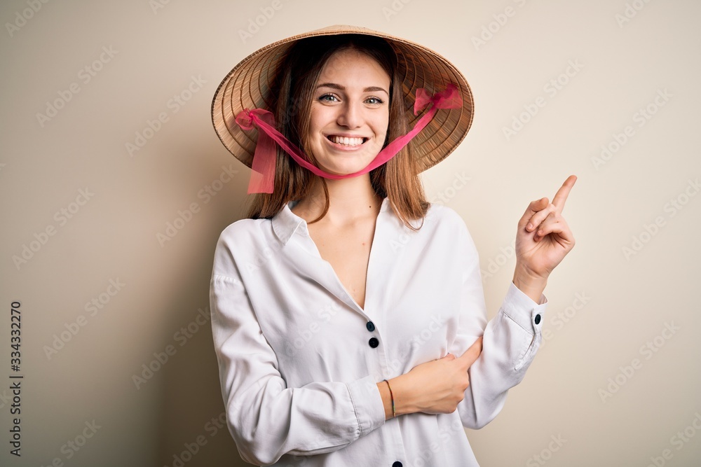 Young beautiful redhead woman wearing asian traditional conical hat over white background with a big smile on face, pointing with hand and finger to the side looking at the camera. Young beautiful redhead woman wearing asian traditional conical hat over white background with a big smile on face, pointing with hand and finger to the side looking at the camera.