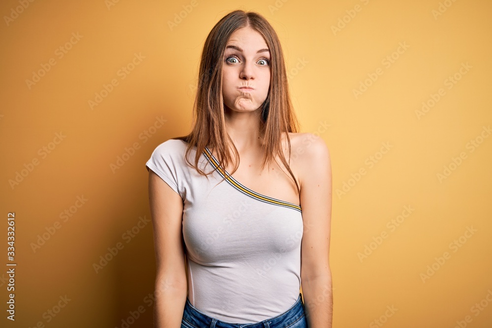 Young beautiful redhead woman wearing casual t-shirt over isolated yellow background puffing cheeks with funny face. Mouth inflated with air, crazy expression. Young beautiful redhead woman wearing casual t-shirt over isolated yellow background puffing cheeks with funny face. Mouth inflated with air, crazy expression.