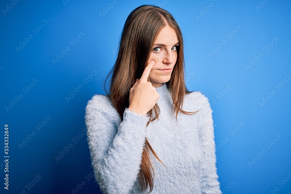 Young beautiful redhead woman wearing casual sweater over isolated blue background Pointing to the eye watching you gesture, suspicious expression Young beautiful redhead woman wearing casual sweater over isolated blue background Pointing to the eye watching you gesture, suspicious expression