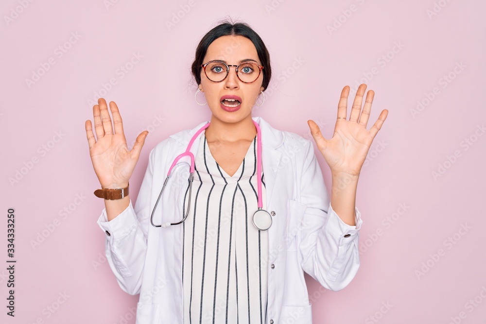 Beautiful doctor woman with blue eyes wearing coat and stethoscope over pink background afraid and terrified with fear expression stop gesture with hands, shouting in shock. Panic concept. Beautiful doctor woman with blue eyes wearing coat and stethoscope over pink background afraid and terrified with fear expression stop gesture with hands, shouting in shock. Panic concept.