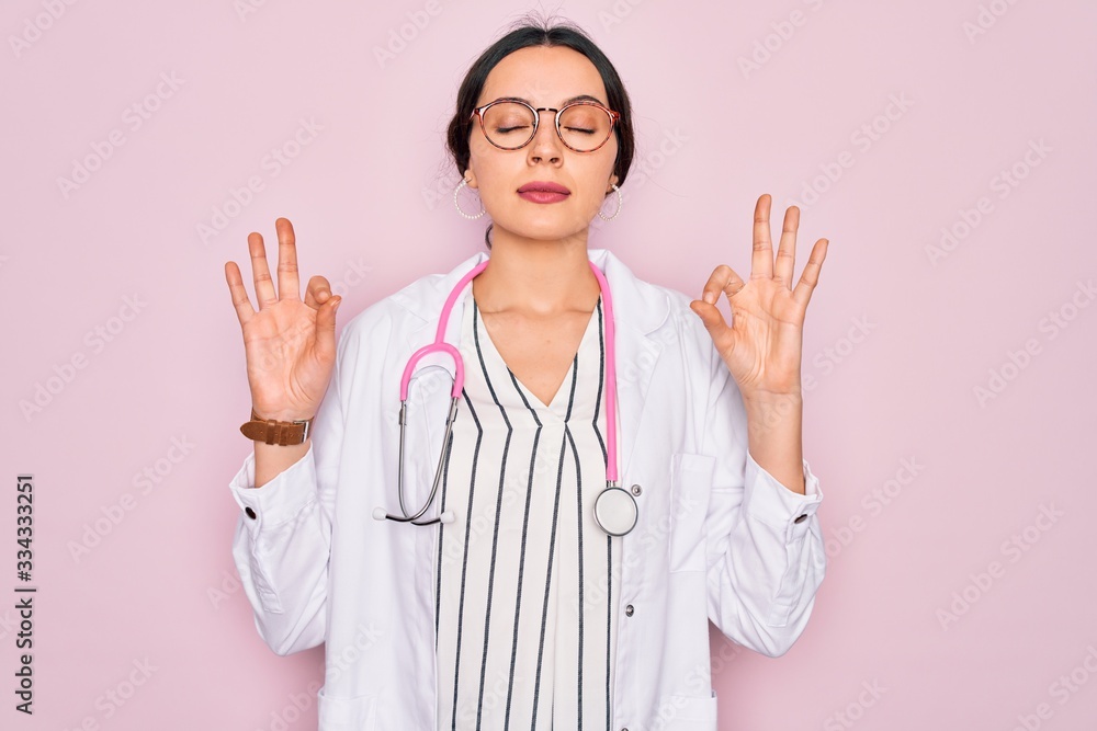 Beautiful doctor woman with blue eyes wearing coat and stethoscope over pink background relax and smiling with eyes closed doing meditation gesture with fingers. Yoga concept. Beautiful doctor woman with blue eyes wearing coat and stethoscope over pink background relax and smiling with eyes closed doing meditation gesture with fingers. Yoga concept.