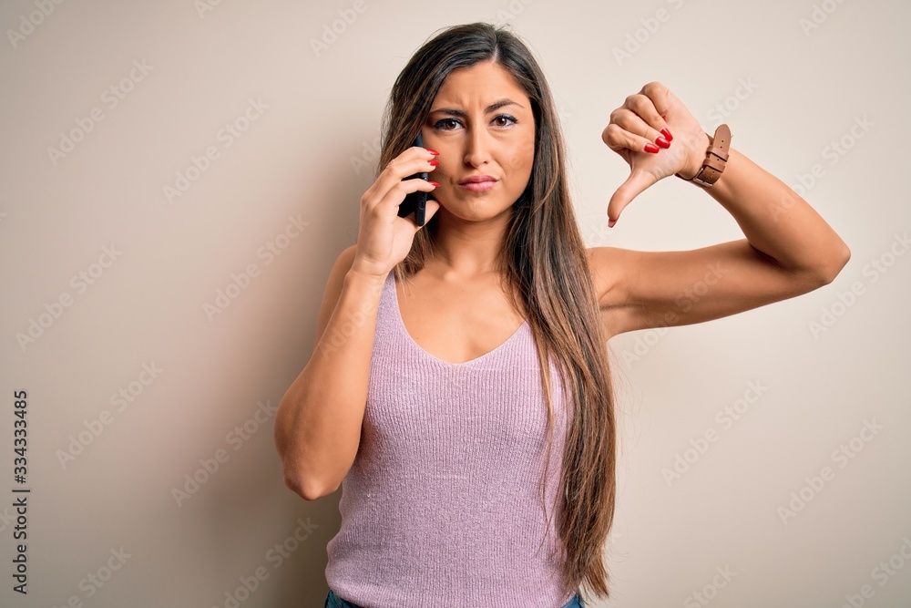 Young brunette woman having conversation talking on the smartphone over white background with angry face, negative sign showing dislike with thumbs down, rejection concept Young brunette woman having conversation talking on the smartphone over white background with angry face, negative sign showing dislike with thumbs down, rejection concept