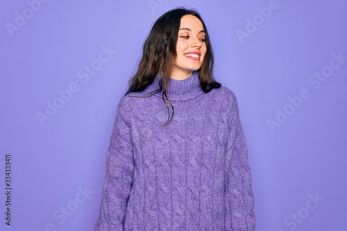Young beautiful woman wearing casual turtleneck sweater standing over purple background looking away to side with smile on face, natural expression. Laughing confident. © Krakenimages.com
