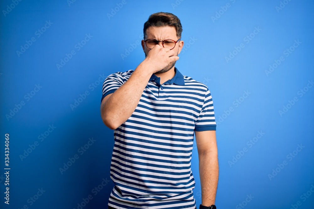Young man with blue eyes wearing glasses and casual striped t-shirt over blue background smelling something stinky and disgusting, intolerable smell, holding breath with fingers on nose. Bad smell