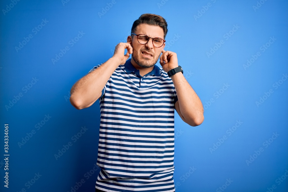 Fototapeta premium Young man with blue eyes wearing glasses and casual striped t-shirt over blue background covering ears with fingers with annoyed expression for the noise of loud music. Deaf concept.