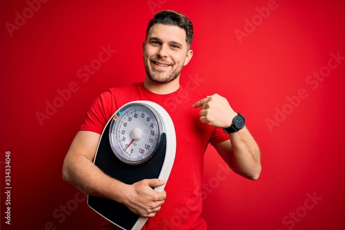 Young fitness man with blue eyes holding scale dieting for healthy weight over red background with surprise face pointing finger to himself