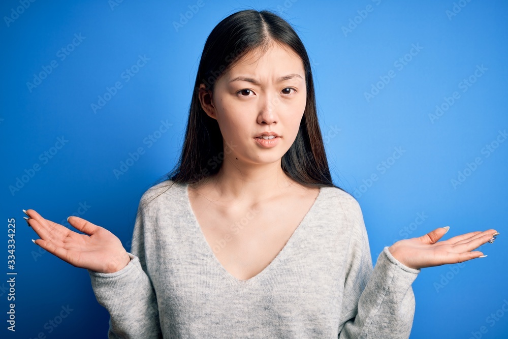 Young beautiful asian woman wearing casual sweater standing over blue isolated background clueless and confused with open arms, no idea concept. Young beautiful asian woman wearing casual sweater standing over blue isolated background clueless and confused with open arms, no idea concept.