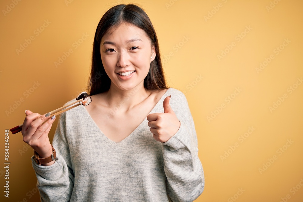 Young asian woman eating japanese food, holding salmon and rice maki sushi using chopsticks happy with big smile doing ok sign, thumb up with fingers, excellent sign Young asian woman eating japanese food, holding salmon and rice maki sushi using chopsticks happy with big smile doing ok sign, thumb up with fingers, excellent sign
