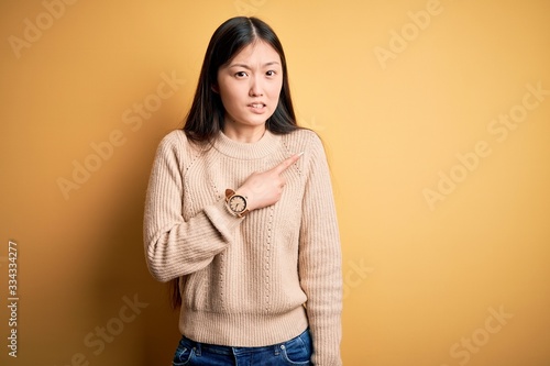 Young beautiful asian woman wearing casual sweater over yellow isolated background Pointing aside worried and nervous with forefinger, concerned and surprised expression