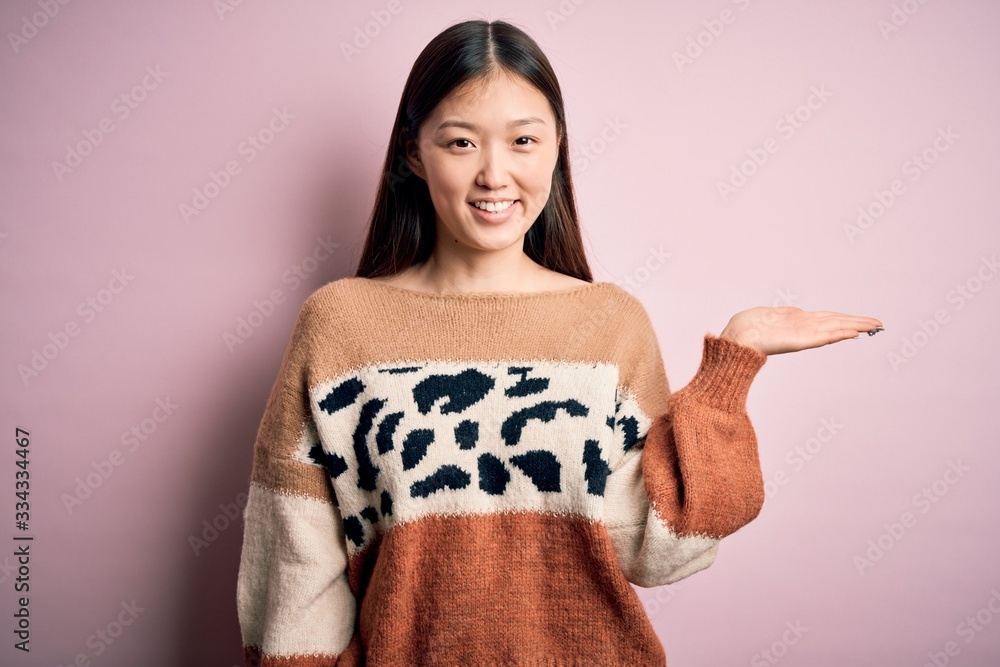 Young beautiful asian woman wearing animal print fashion sweater over pink isolated background smiling cheerful presenting and pointing with palm of hand looking at the camera. Young beautiful asian woman wearing animal print fashion sweater over pink isolated background smiling cheerful presenting and pointing with palm of hand looking at the camera.