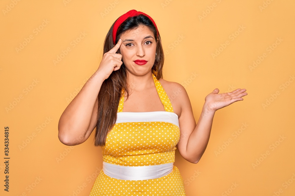 Young hispanic pin up woman wearing fashion sexy 50s style over yellow background confused and annoyed with open palm showing copy space and pointing finger to forehead. Think about it.