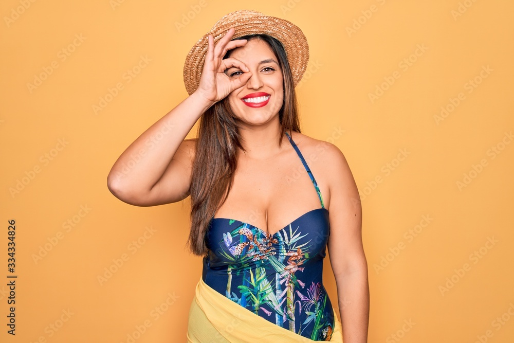 Young hispanic woman wearing summer hat and swimsuit over yellow background doing ok gesture with hand smiling, eye looking through fingers with happy face. Young hispanic woman wearing summer hat and swimsuit over yellow background doing ok gesture with hand smiling, eye looking through fingers with happy face.
