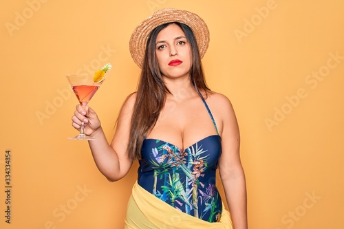 Young hispanic woman wearing summer swimsuit drinking a cocktail over yellow background with a confident expression on smart face thinking serious