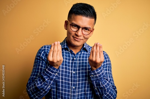 Young handsome latin man wearing casual shirt and glasses over yellow background doing money gesture with hands, asking for salary payment, millionaire business