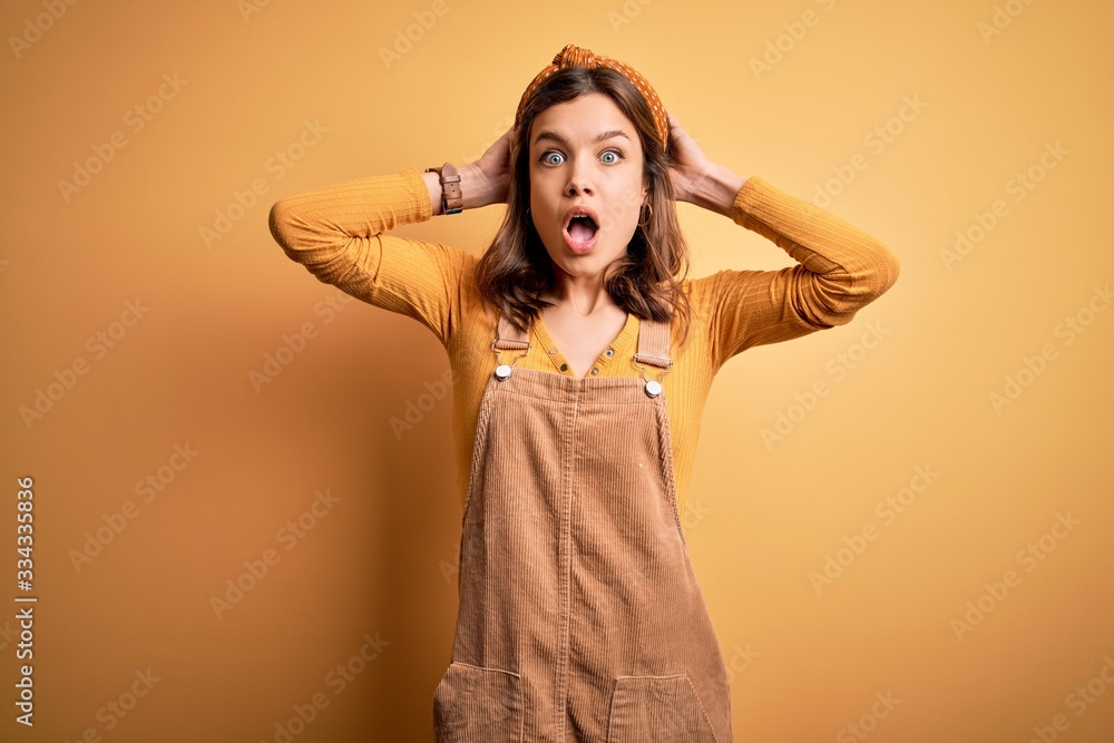 Fototapeta premium Young beautiful blonde girl wearing overall standing over yellow isolated background Crazy and scared with hands on head, afraid and surprised of shock with open mouth