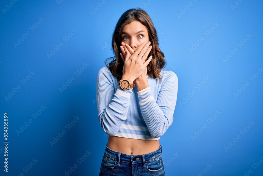 Young beautiful blonde girl wearing casual sweater standing over blue isolated background shocked covering mouth with hands for mistake. Secret concept. Young beautiful blonde girl wearing casual sweater standing over blue isolated background shocked covering mouth with hands for mistake. Secret concept.