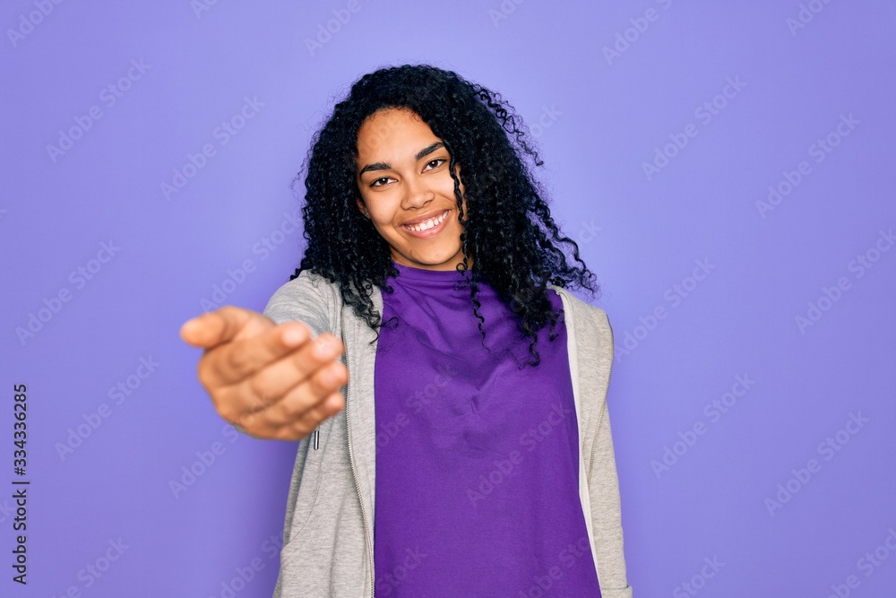 Young african american sporty woman wearing casual sweatshirt over purple background smiling friendly offering handshake as greeting and welcoming. Successful business. Young african american sporty woman wearing casual sweatshirt over purple background smiling friendly offering handshake as greeting and welcoming. Successful business.