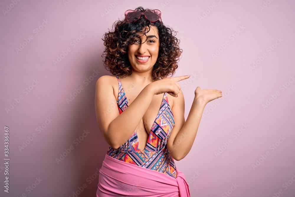 Young beautiful arab woman on vacation wearing swimsuit and sunglasses over pink background amazed and smiling to the camera while presenting with hand and pointing with finger. Young beautiful arab woman on vacation wearing swimsuit and sunglasses over pink background amazed and smiling to the camera while presenting with hand and pointing with finger.