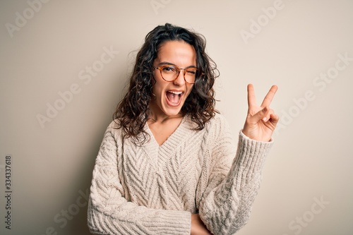 Beautiful woman with curly hair wearing casual sweater and glasses over white background smiling with happy face winking at the camera doing victory sign. Number two.