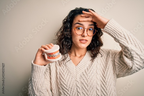 Young beautiful woman with curly hair holding plastic teeth with dental baces stressed with hand on head, shocked with shame and surprise face, angry and frustrated. Fear and upset for mistake. photo