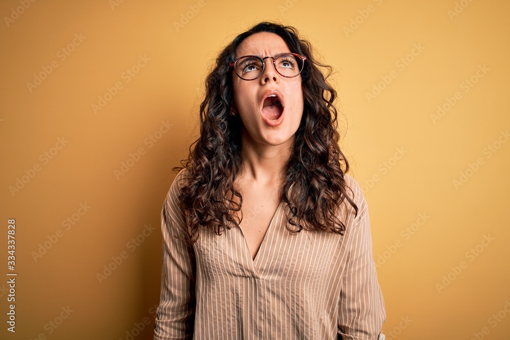 Beautiful woman with curly hair wearing striped shirt and glasses over yellow background angry and mad screaming frustrated and furious, shouting with anger. Rage and aggressive concept. Beautiful woman with curly hair wearing striped shirt and glasses over yellow background angry and mad screaming frustrated and furious, shouting with anger. Rage and aggressive concept.
