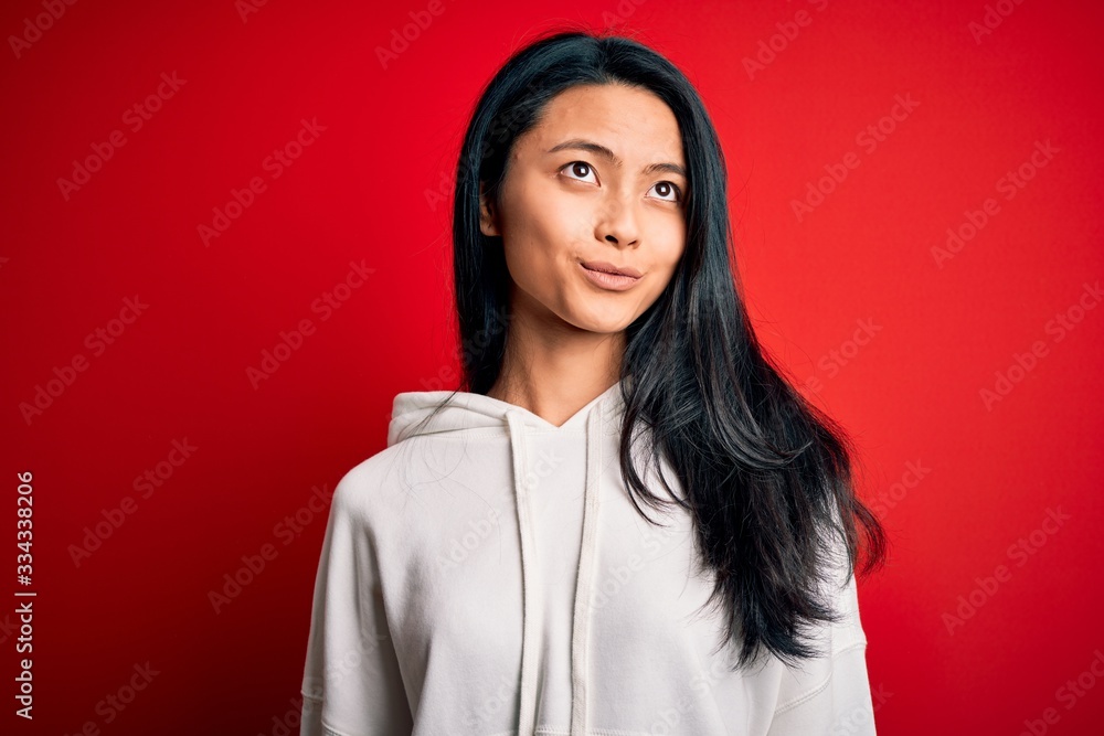 Young beautiful chinese sporty woman wearing sweatshirt over isolated red background smiling looking to the side and staring away thinking. Young beautiful chinese sporty woman wearing sweatshirt over isolated red background smiling looking to the side and staring away thinking.