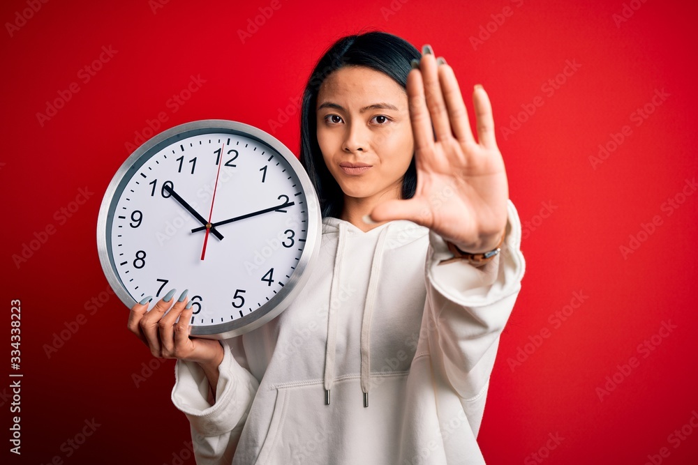 Young beautiful chinese woman holding big clock standing over isolated red background with open hand doing stop sign with serious and confident expression, defense gesture Young beautiful chinese woman holding big clock standing over isolated red background with open hand doing stop sign with serious and confident expression, defense gesture