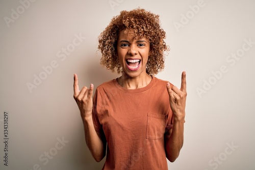 Beautiful african american woman with curly hair wearing casual t-shirt over white background shouting with crazy expression doing rock symbol with hands up. Music star. Heavy concept.
