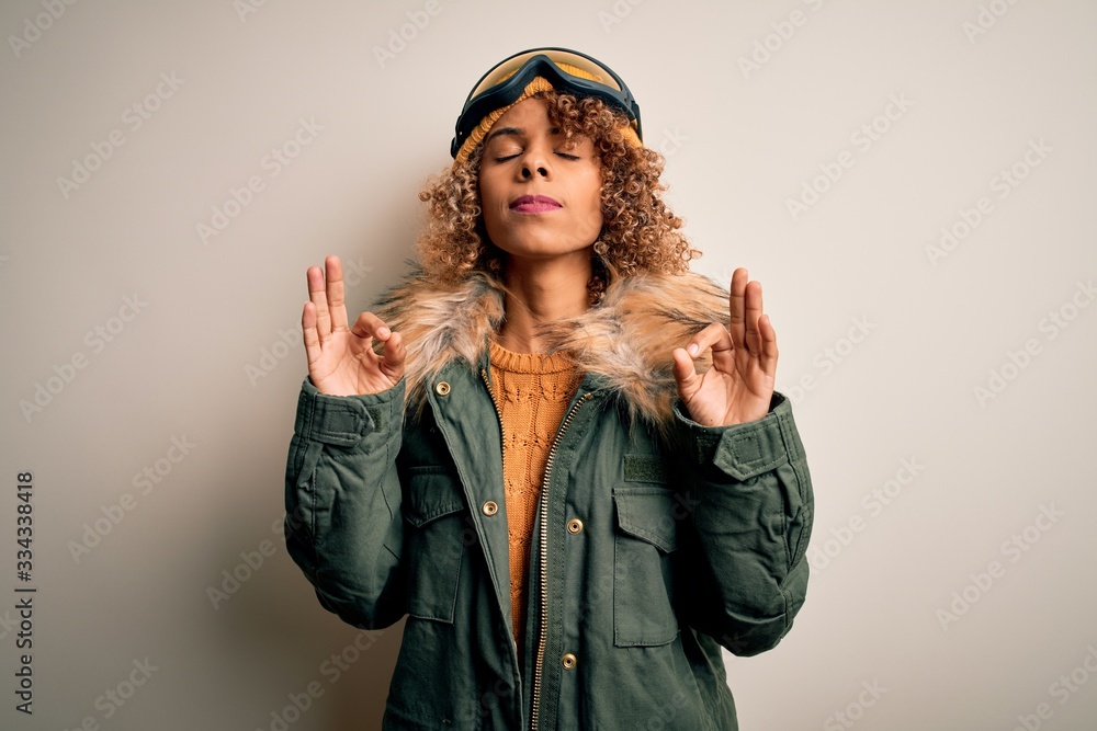Young african american skier woman with curly hair wearing snow sportswear and ski goggles relaxed and smiling with eyes closed doing meditation gesture with fingers. Yoga concept.
