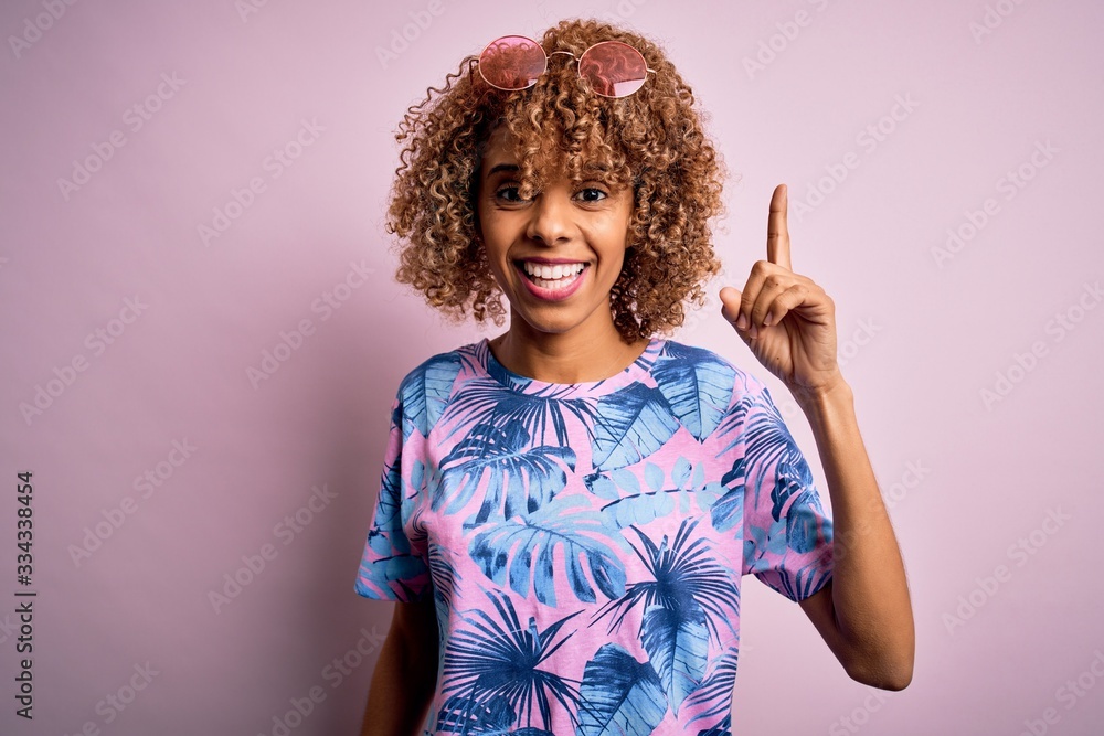 Young african american woman on vacation wearing summer t-shirt over pink background pointing finger up with successful idea. Exited and happy. Number one. Young african american woman on vacation wearing summer t-shirt over pink background pointing finger up with successful idea. Exited and happy. Number one.