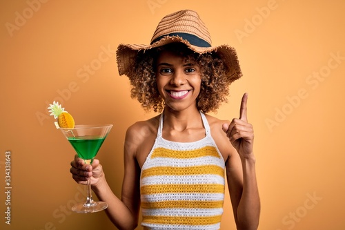 African american tourist woman with curly on vacation wearing summer hat drinking cocktail surprised with an idea or question pointing finger with happy face, number one