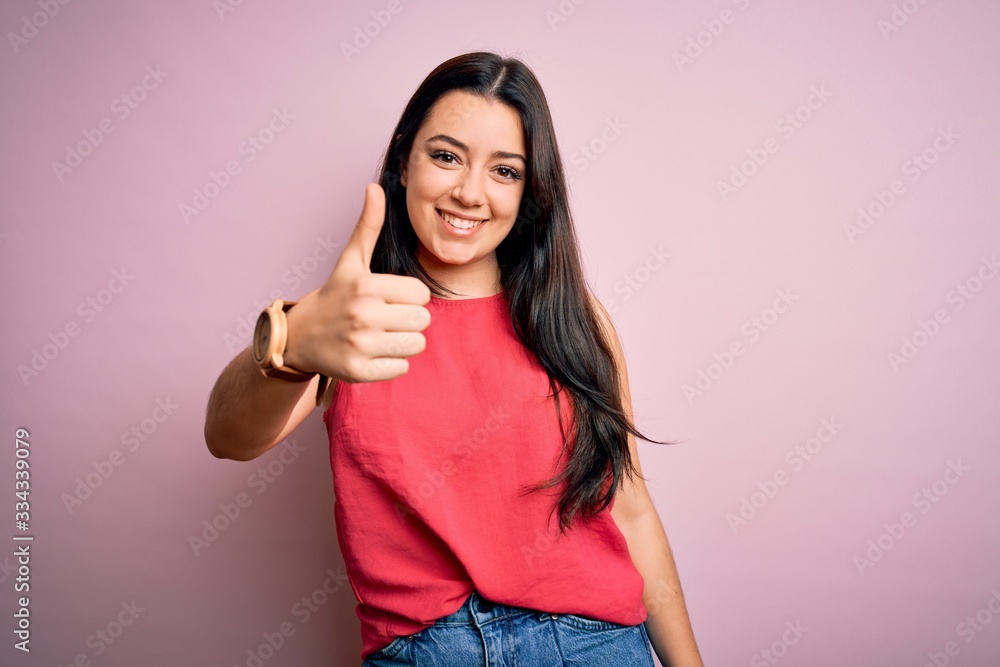 Young brunette woman wearing casual summer shirt over pink isolated background doing happy thumbs up gesture with hand. Approving expression looking at the camera showing success. Young brunette woman wearing casual summer shirt over pink isolated background doing happy thumbs up gesture with hand. Approving expression looking at the camera showing success.