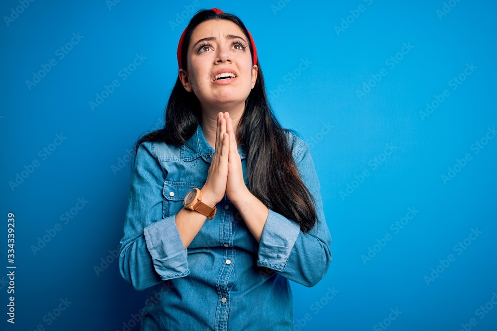 Young brunette woman wearing casual denim shirt over blue isolated background begging and praying with hands together with hope expression on face very emotional and worried. Begging. Young brunette woman wearing casual denim shirt over blue isolated background begging and praying with hands together with hope expression on face very emotional and worried. Begging.
