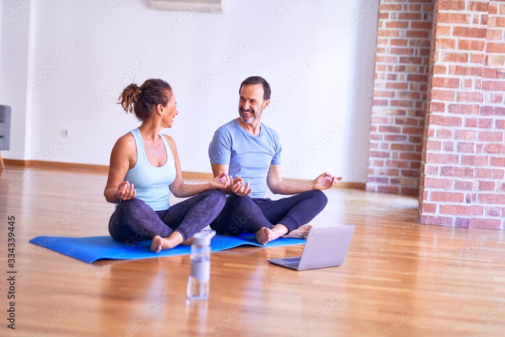 Middle age beautiful sporty couple smiling happy. Sitting on mat practicing yoga doing lotus pose using laptop at gym