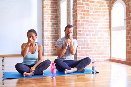 Middle age sporty couple sitting on mat doing stretching yoga exercise at gym smelling something stinky and disgusting  intolerable smell  holding breath with fingers on nose. Bad smells concept.