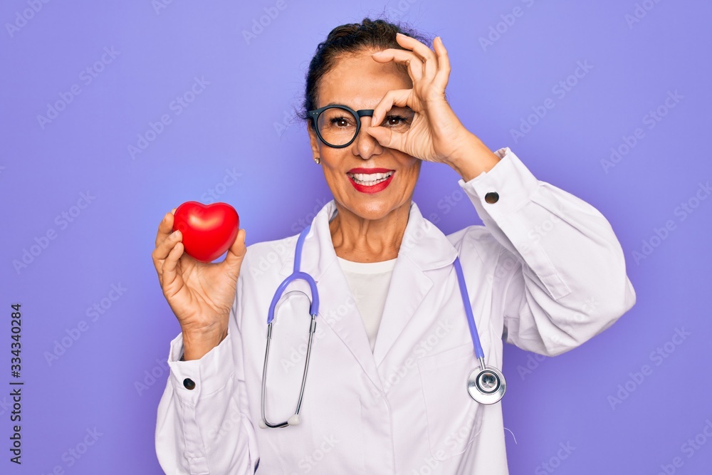 Middle age senior cardiologist doctor woman holding red heart over purple background with happy face smiling doing ok sign with hand on eye looking through fingers