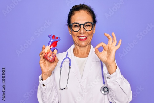 Middle age senior cardiologist doctor woman holding professional cardiology heart doing ok sign with fingers, excellent symbol photo