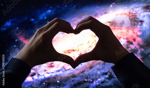 close up to a two male hands making sign heart by fingers to a beautiful colorful circular galaxy at background. Love Universe concept and science concept