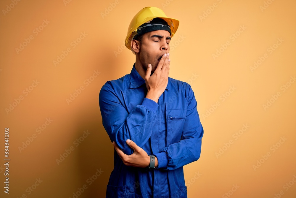 Young handsome african american worker man wearing blue uniform and security helmet bored yawning tired covering mouth with hand. Restless and sleepiness.