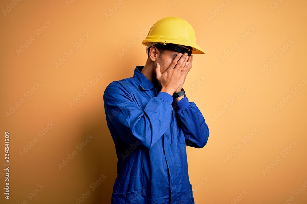 Young handsome african american worker man wearing blue uniform and security helmet with sad expression covering face with hands while crying. Depression concept.