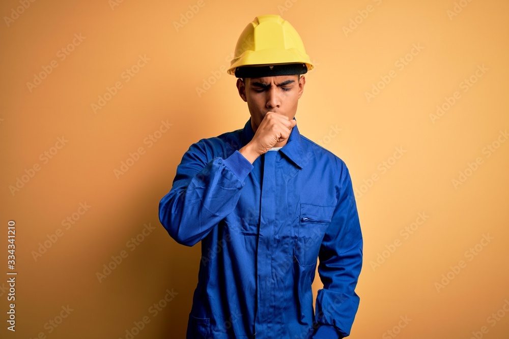 Young handsome african american worker man wearing blue uniform and security helmet feeling unwell and coughing as symptom for cold or bronchitis. Health care concept. Young handsome african american worker man wearing blue uniform and security helmet feeling unwell and coughing as symptom for cold or bronchitis. Health care concept.