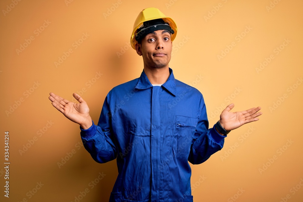 Young handsome african american worker man wearing blue uniform and security helmet clueless and confused expression with arms and hands raised. Doubt concept.