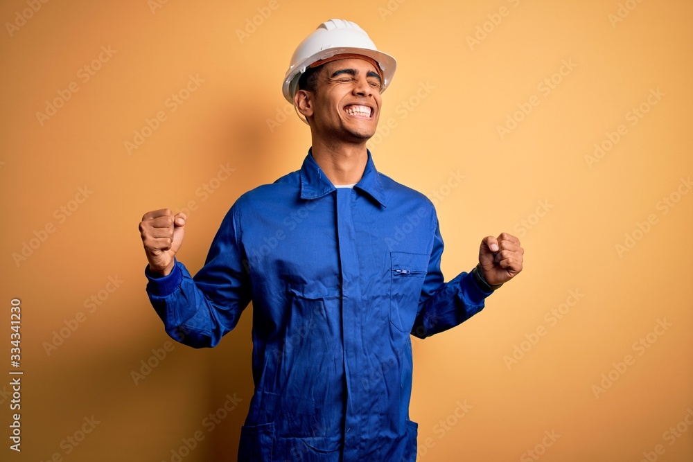 Young handsome african american worker man wearing blue uniform and security helmet very happy and excited doing winner gesture with arms raised, smiling and screaming for success. Celebration