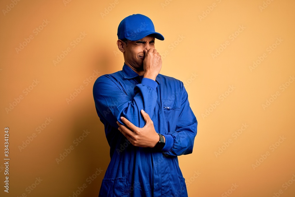 Young african american mechanic man wearing blue uniform and cap over yellow background smelling something stinky and disgusting, intolerable smell, holding breath with fingers on nose. Bad smell