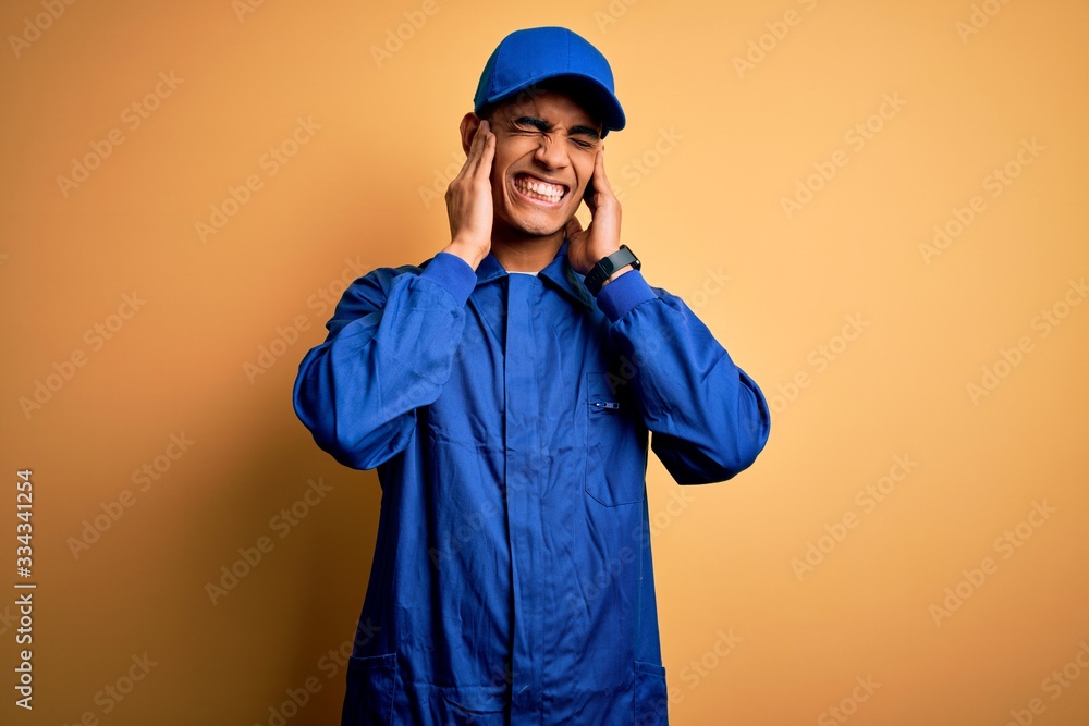 Young african american mechanic man wearing blue uniform and cap over yellow background covering ears with fingers with annoyed expression for the noise of loud music. Deaf concept.
