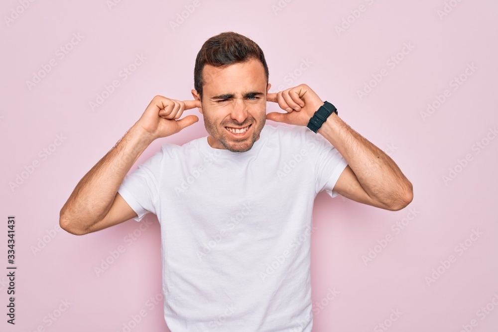 Handsome man with blue eyes wearing casual white t-shirt standing over pink background covering ears with fingers with annoyed expression for the noise of loud music. Deaf concept. Handsome man with blue eyes wearing casual white t-shirt standing over pink background covering ears with fingers with annoyed expression for the noise of loud music. Deaf concept.