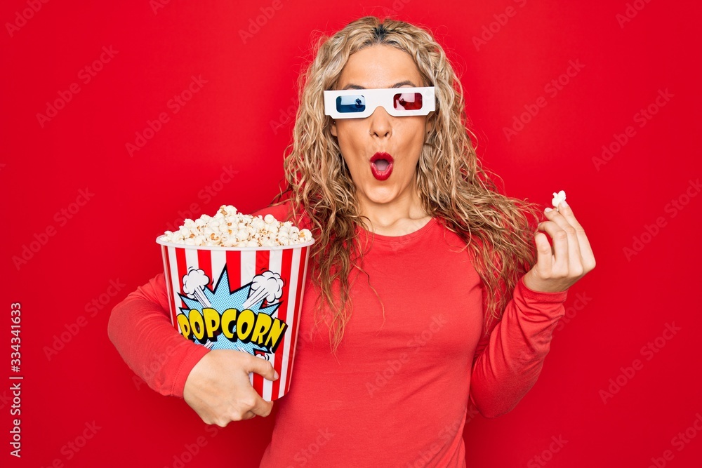 Obraz premium Young beautiful blonde woman watching movie using 3d glasses eating popcorn scared and amazed with open mouth for surprise, disbelief face